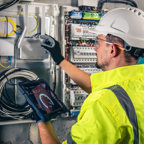 Man, an electrical technician working in a switchboard with fuses. Installation and connection of electrical equipment. Professional uses a tablet.